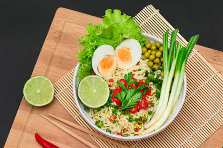 Beautiful Noodle Dish with Green Pea, Eggs, Red Hot Pepper, Greens and Lime with Chopsticks on Bamboo Cutting Board. Instant Noodles with Vegetables - Top Viewの写真素材