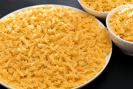 Uncooked Fusilli Pasta Lying on White Plate on Black Background. Raw and Dry Macaroni. Unhealthy and Fat Foodの写真素材