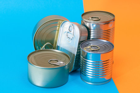 A Group of Stacked Tin Cans with Blank Edges on Split Blue and Orange Background. Canned Food. Different Aluminum Cans for Safe and Long Term Storage of Food. Steel Sealed Food Storage Containersの写真素材