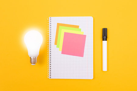 Glowing Power Saving Lamp and Notepad with Sticky Notes Lying on Yellow Table, Blank Template with Copy Space - Top View, Flat Layの写真素材