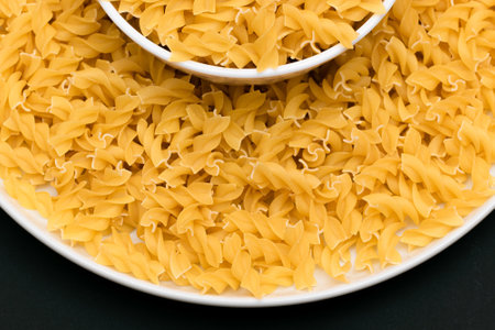 Uncooked Fusilli Pasta Lying on White Plate on Black Background. Raw and Dry Macaroni. Unhealthy and Fat Food - Top Viewの写真素材