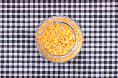 Uncooked Chifferi Rigati Pasta in Glass Jar on Black Checkered Towel. Fat and Unhealthy Food. Scattered Classic Dry Macaroni. Italian Culture and Cuisine. Raw Pastaの写真素材