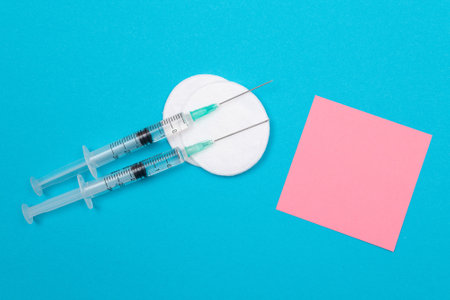 Vaccination, Immunology or Revaccination Concept - Two Medical Disposable Syringe Lying on Blue Table in Doctors Office in a Hospital or Clinic. Blank Pink Sticky Note - Mock Up with Copy Spaceの写真素材