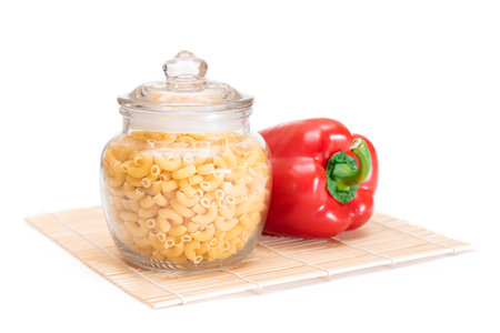 Uncooked Chifferi Rigati Pasta in Glass Jar and Red Bell Pepper on Bamboo Mat Isolated on White Background. Fat and Unhealthy Food. Classic Dry Macaroni. Italian Culture and Cuisine. Raw Pastaの写真素材
