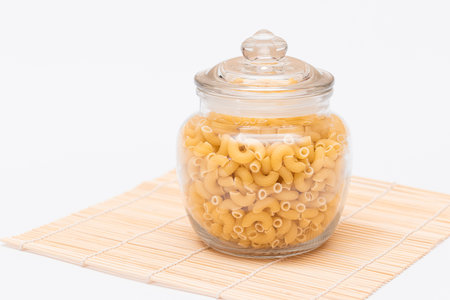 Uncooked Chifferi Rigati Pasta in Glass Jar on Bamboo Mat on White Background. Fat and Unhealthy Food. Classic Dry Macaroni. Italian Culture and Cuisine. Raw Pastaの写真素材