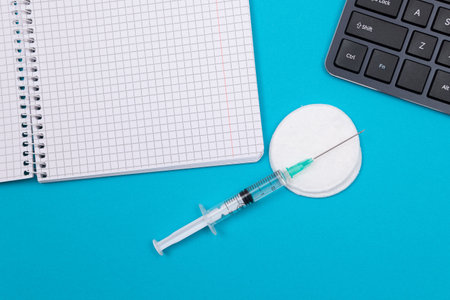 Vaccination, Immunology or Revaccination Concept - A Medical Syringe Lying on Blue Table in Doctors Office in a Hospital or Clinic. Opened Blank Notepad - Mock Up with Copy Spaceの写真素材