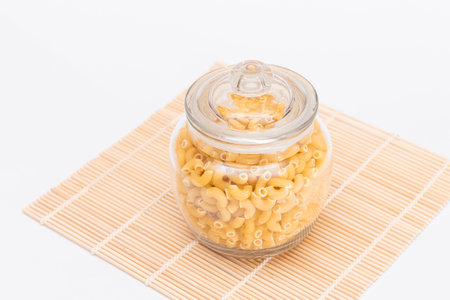 Uncooked Chifferi Rigati Pasta in Glass Jar on Bamboo Mat on White Background. Fat and Unhealthy Food. Classic Dry Macaroni. Italian Culture and Cuisine. Raw Pastaの写真素材