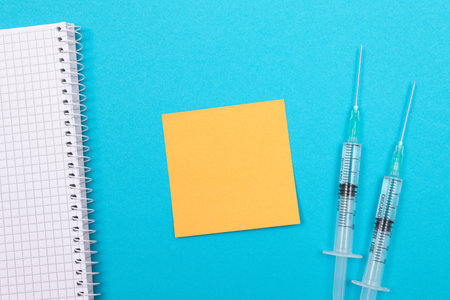 Vaccination, Immunology or Revaccination Concept - Two Medical Disposable Syringe Lying on Blue Table in Doctors Office in a Hospital or Clinic. Blank Orange Sticky Note - Mock Up with Copy Spaceの写真素材