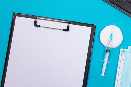 Vaccination, Immunology or Revaccination Concept - A Medical Syringe Lying on Blue Table in Doctors Office in a Hospital or Clinic. Black Clipboard with Sheet of Paper - Mock Up with Copy Spaceの写真素材