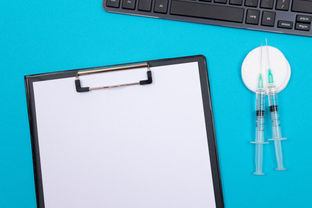 Vaccination, Immunology or Revaccination Concept - Two Medical Syringe Lying on Blue Table in Doctors Office in a Hospital or Clinic. Black Clipboard with Sheet of Paper - Mock Up with Copy Spaceの写真素材