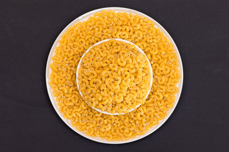Uncooked Chifferi Rigati Pasta in White Plate on Black Background. Fat and Unhealthy Food. Scattered Classic Dry Macaroni. Italian Culture and Cuisine. Raw Pastaの写真素材