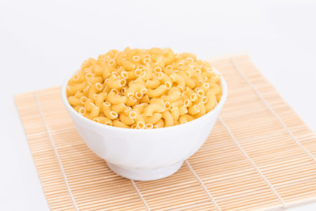 Uncooked Chifferi Rigati Pasta in White Cup on Bamboo Mat on White Background. Fat and Unhealthy Food. Scattered Classic Dry Macaroni. Italian Culture and Cuisine. Raw Pastaの写真素材
