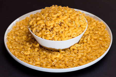 Uncooked Chifferi Rigati Pasta in White Plate on Black Background. Fat and Unhealthy Food. Scattered Classic Dry Macaroni. Italian Culture and Cuisine. Raw Pastaの写真素材