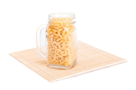 Uncooked Chifferi Rigati Pasta in Glass Jar on Bamboo Mat Isolated on White Background. Fat and Unhealthy Food. Scattered Classic Dry Macaroni. Italian Culture and Cuisine. Raw Pasta - Isolationの写真素材