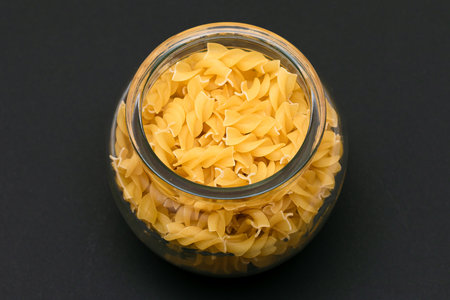 Uncooked Fusilli Pasta in Glass Jar on Black Background. Raw and Dry Macaroni. Unhealthy and Fat Food. Italian Cultureの写真素材