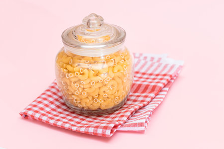Uncooked Chifferi Rigati Pasta in Glass Jar on Checkered Red Towel on Pink Background. Fat and...