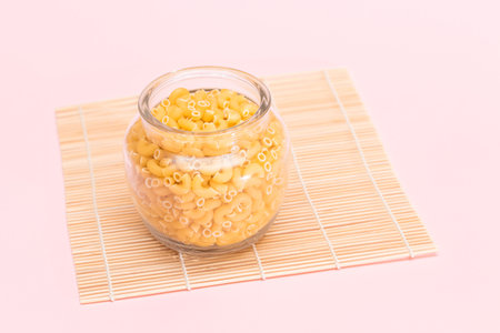 Uncooked Chifferi Rigati Pasta in Glass Jar on Bamboo Mat on Pink Background. Fat and Unhealthy Food. Scattered Classic Dry Macaroni. Italian Culture and Cuisine. Raw Pastaの写真素材