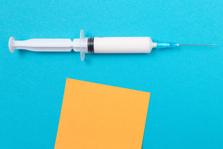 Vaccination, Immunology or Revaccination Concept - A Medical Disposable Syringe Lying on Blue Table in Doctors Office in a Hospital or Clinic. Blank Orange Sticky Note - Mock Up with Copy Spaceの写真素材