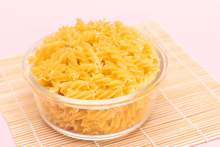 Uncooked Fusilli Pasta in Glass Jar on Bambo Mat on Pink Background. Fat and Unhealthy Food. Classic Dry Spiral Macaroni. Italian Culture and Cuisine. Raw Pastaの写真素材