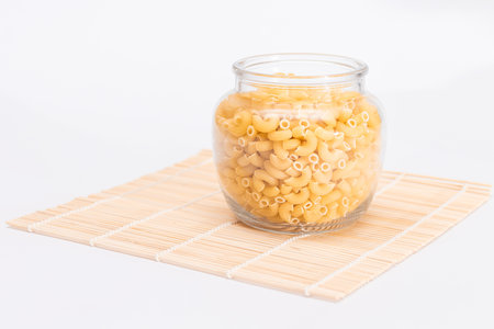 Uncooked Chifferi Rigati Pasta in Glass Jar on Bamboo Mat on White Background. Fat and Unhealthy Food. Classic Dry Macaroni. Italian Culture and Cuisine. Raw Pastaの写真素材