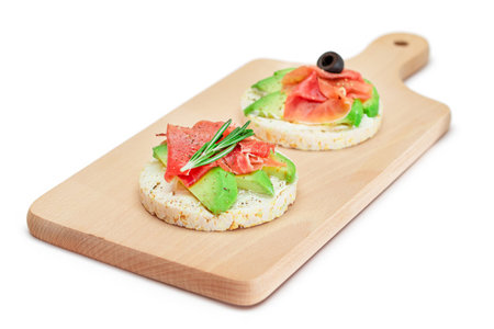 Rice Cake Sandwich with Avocado, Jamon, Olives and Rosemary on Wooden Cutting Board - Isolated on White. Easy Breakfast. Quick and Healthy Sandwiches. Crispbread with Tasty Filling - Isolationの写真素材