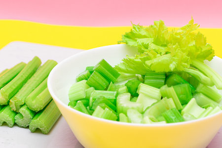 Fresh Chopped Celery Slices in White Bowl with Celery Sticks on White Cutting Board. Vegan and Vegetarian Culture. Raw Food. Healthy Diet with Negative Calorie Contentの写真素材
