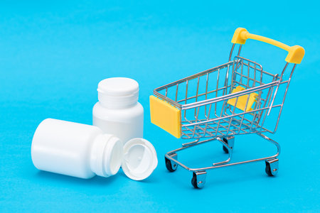 Expensive Medicine and Inflation: Scattered Pills or Capsules and Shopping Cart on Blue Background. Global Pharmaceutical Industry and Big Pharma. Ordering Pharmaceutical Productsの写真素材