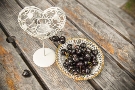 fresh red cherries berries in a white plate on the wooden table with candlestick in the shape of the heart outdoors in the garden. Romantic place.の写真素材