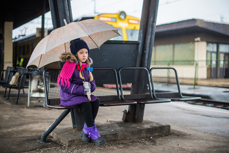 Little child sitting on platform with an umbrella on a railway train station. Rainy weather. Kid girl waiting for train. Child lost.の写真素材