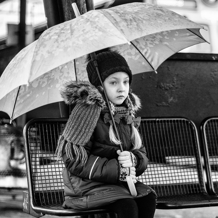 Little child sitting on platform with an umbrella on a railway train station. Rainy weather. Kid girl waiting for train. Child lost.の写真素材
