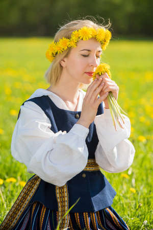 Young woman in national clothes wearing yellow dandelion wreath in spring field. Springtimeの写真素材