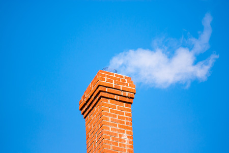 Red brick chimney releasing white smoke on clear blue sky の写真素材