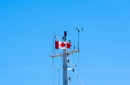 Top of modern metal ship mast against empty blue sky copy space, with antennas, rigging, weather vane, and Canadian flag fluttering in wind.の写真素材