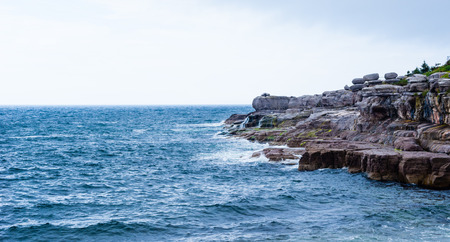Rocky coastline with ocean waves splashing on shore against overcast sky, in Newfoundland, Canada.