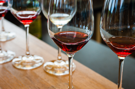 Detail of wine glasses with red wine samples, on wood counter with other glasses in background.