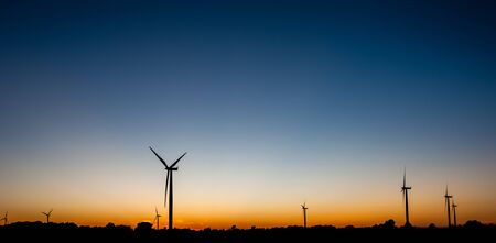Black silhouettes of several wind turbines against orange and blue sky at dusk.の写真素材