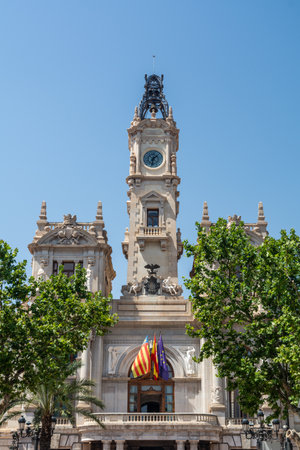Detailed view of the ornate clock tower, sculptural reliefs and flag-draped balcony of Valencia City Hall framed by leafy plane trees.の写真素材