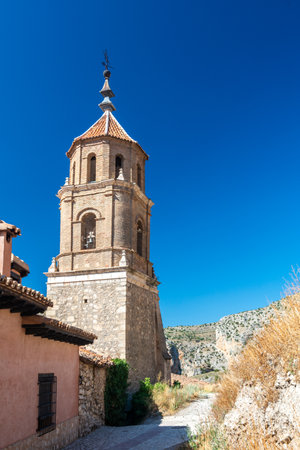 Iglesia de Santiago Apostol bell tower rises above medieval stone walls, backed by rugged limestone hills and a clear blue sky.の写真素材