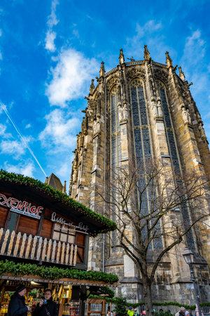 Aachen, Germany - Dec 04, 2025: Aachen Cathedral Gothic choir rises above a Christmas market stall under blue sky as visitors walk through the square.のeditorial素材