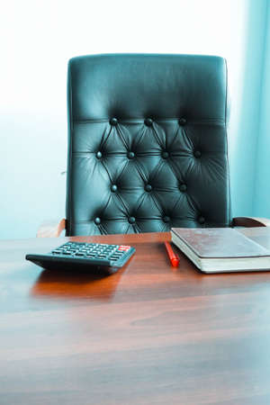 Photo of a calculator, pen and notebook on a wooden brown table near a black leather chair in the...