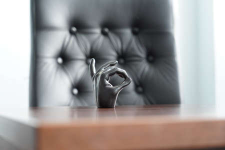 Photo of a black palm hand showing an okay sign stands on a wooden brown table near a black leather chair in the office.の写真素材