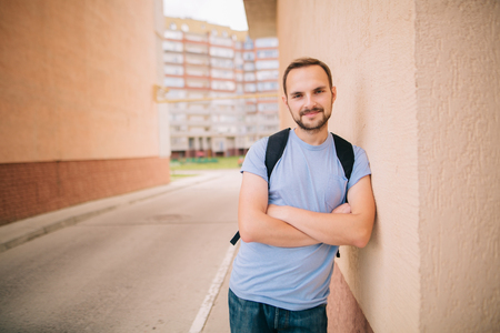 Smiling caucasian bearded student young man hipster in blue t-shirt with backpack standing in arch leaning on wall in pastel beige tones near new building with his arms crossed closeup view.の写真素材