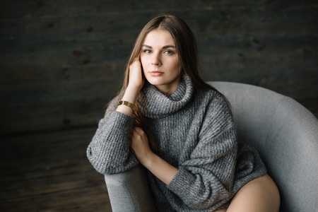 Dark haired sad woman in long grey knitted cozy sweater sitting in a chair at home near big window. Happy and Healthy girl with long beautiful legs with soft skin in studio.の写真素材