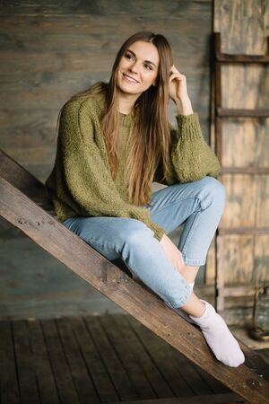 Grunge fashion: cute young girl informal model in blue jeans, dark knitted sweater and white socks sitting on the ladder smiling on the wooden backgroundの写真素材