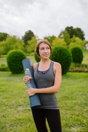 A young female fitness trainer stands in nature and holds a bottle of water and a yoga mat. Outdoor sports, high quality shot.の写真素材