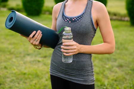 Close-up of a female yoga instructor with a fitness mat and a bottle of water in a gray tank top against a green lawn. High quality shot.の写真素材