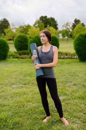 Athletic thin young woman holds yoga mat and bottle of water after or before training outdoors in the park cheerful with smile on face with happy and natural expression in sportswear. Vertical photo.の写真素材