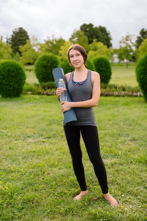 A young athletic girl in a gray tracksuit for fitness is going to do yoga in a green park, in her hands a bottle of water and a yoga mat. High quality photo.の写真素材
