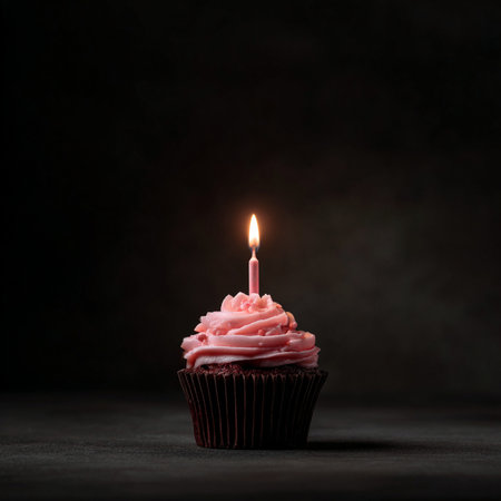 A single cupcake topped with smooth pink frosting and a lit birthday candle, set against a dramatic dark background. This minimal, elegant composition is ideal for birthday invitations, celebration ads, greeting cards, or holiday-themed graphics. The image emphasizes warmth, festivity, and simplicity with a strong visual impactの写真素材