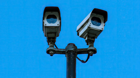 High-resolution image of two surveillance cameras on a pole with a backdrop of a clear blue sky, focusing on their role in urban surveillance.の素材
